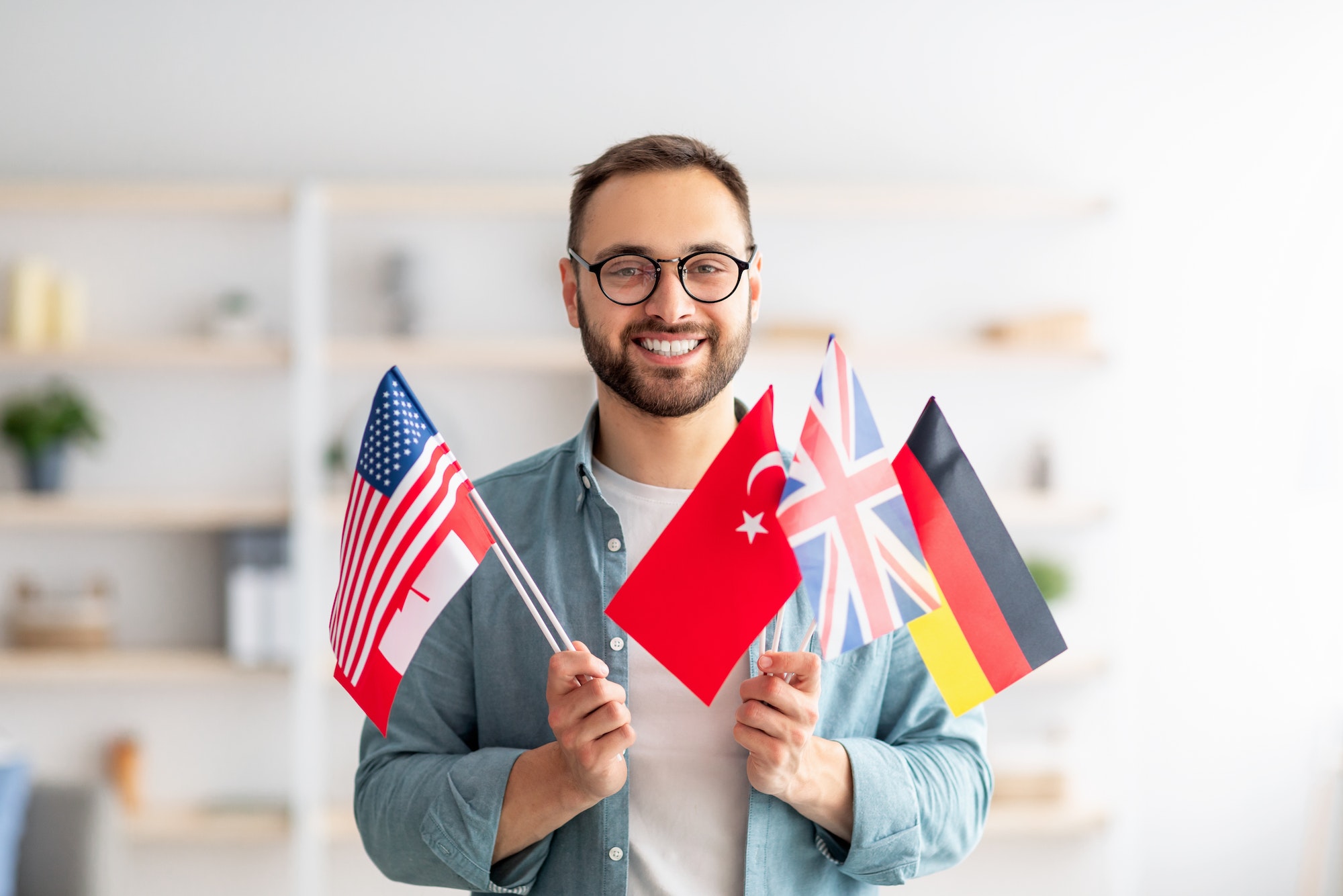 Handsome young guy holding bunch of diverse flags and smiling at camera indoors