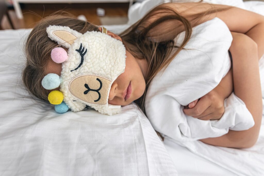 A little girl sleeps on a pillow wearing a funny sleep mask.