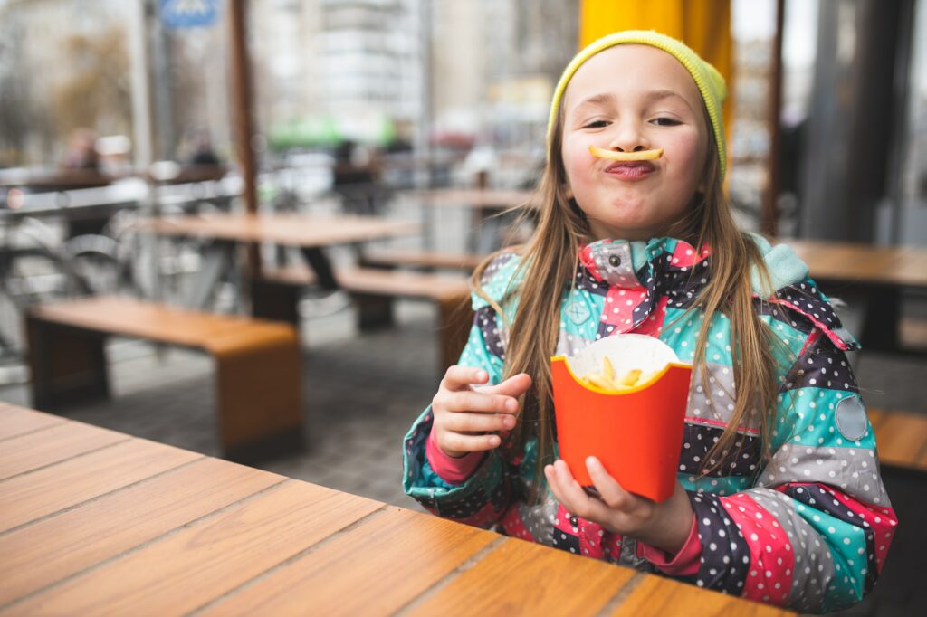 The child eats french fries on the street. The child eats fast food. Funny child eats food.