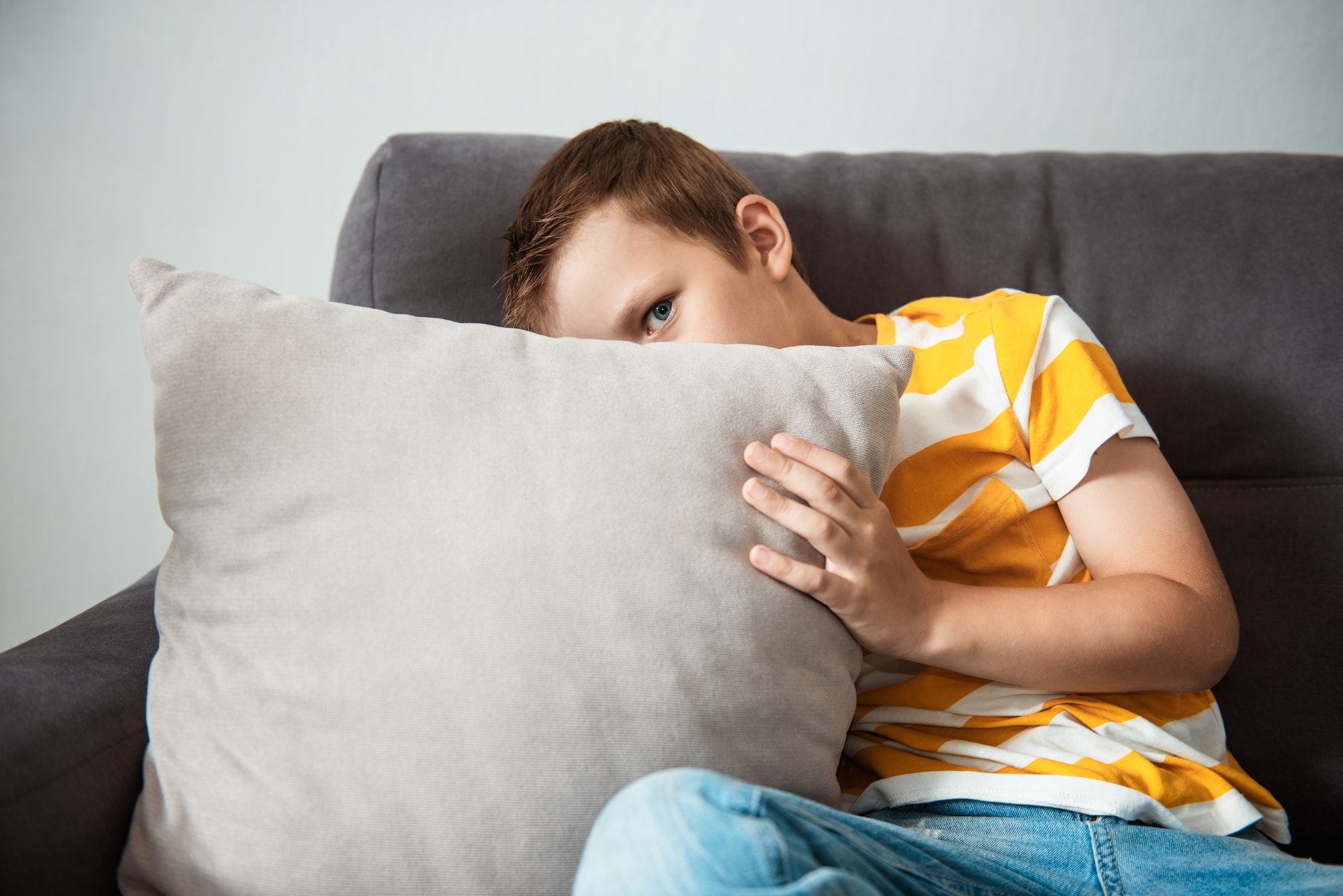Depression in adolescence - a sad child boy sitting on sofa, hugs a pillow with her hands and hides